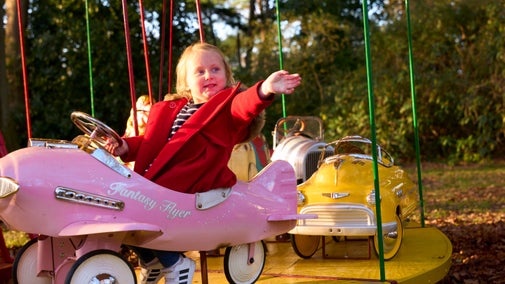 A toddler in a red coat waves whilst riding a pink airplane on a carousel.